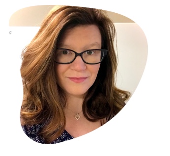 Woman wearing a blue printed shirt and blakc glasses, witha gold heart necklace. Her hair is a light brown with highlight and fair skin. 