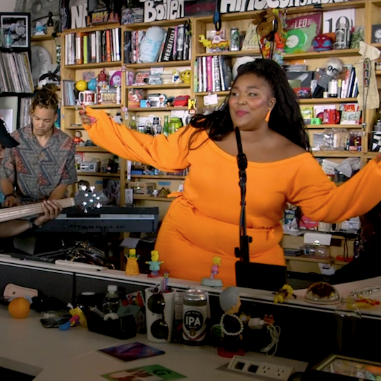 Lizzo an orange dress with her arms wide open with a background of a tiny desk music studio 